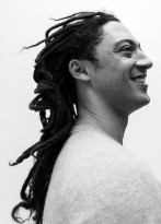 Black and white portrait of a mixed race man with long dreads. He is smiling and standing in profle facing towards the right.